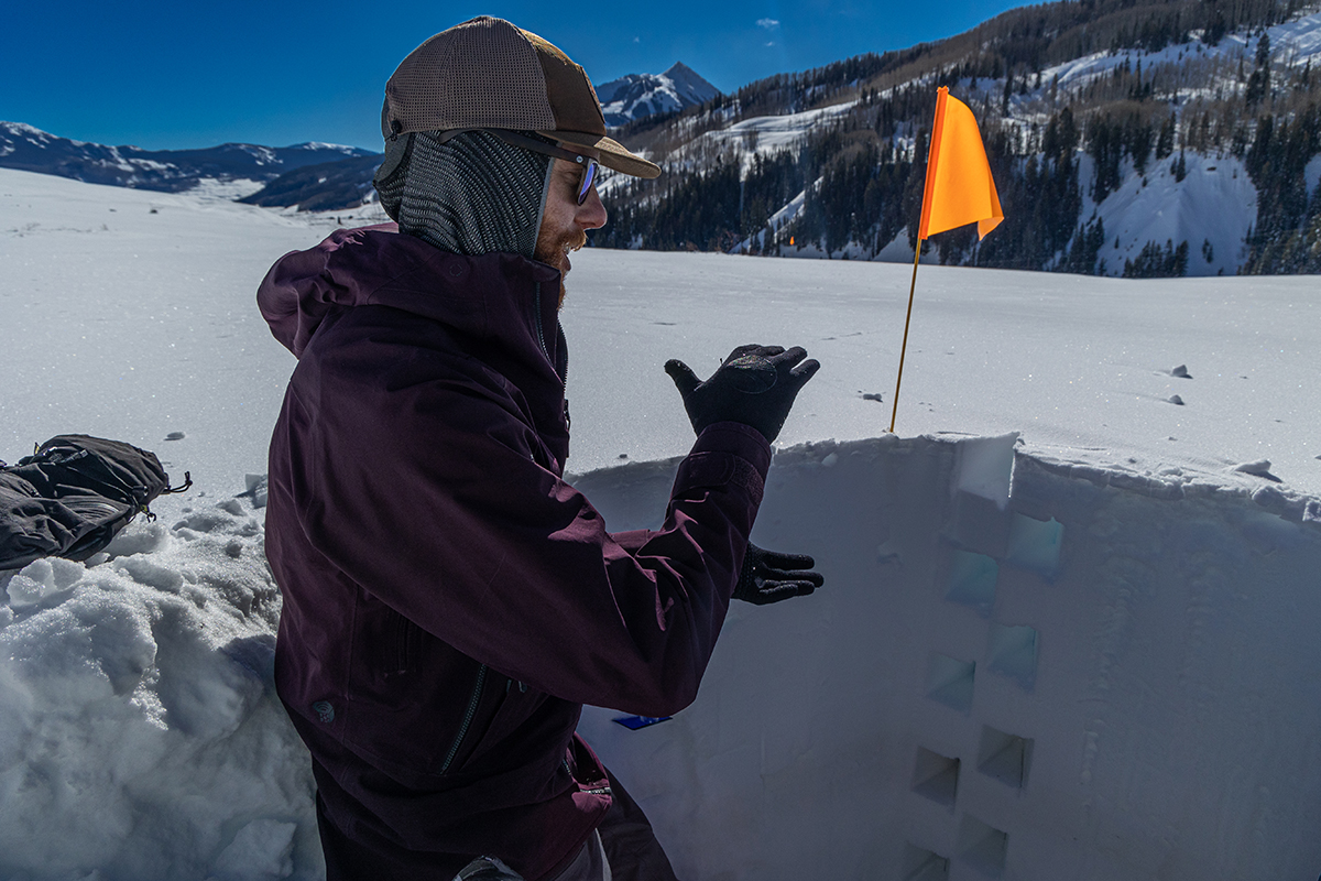 Hogan, a University of Washington PhD student, gestures while describing measurements within a neatly dug snow pit. The square-shaped cutouts indicate where snow samples were removed in triangular prisms to measure density.