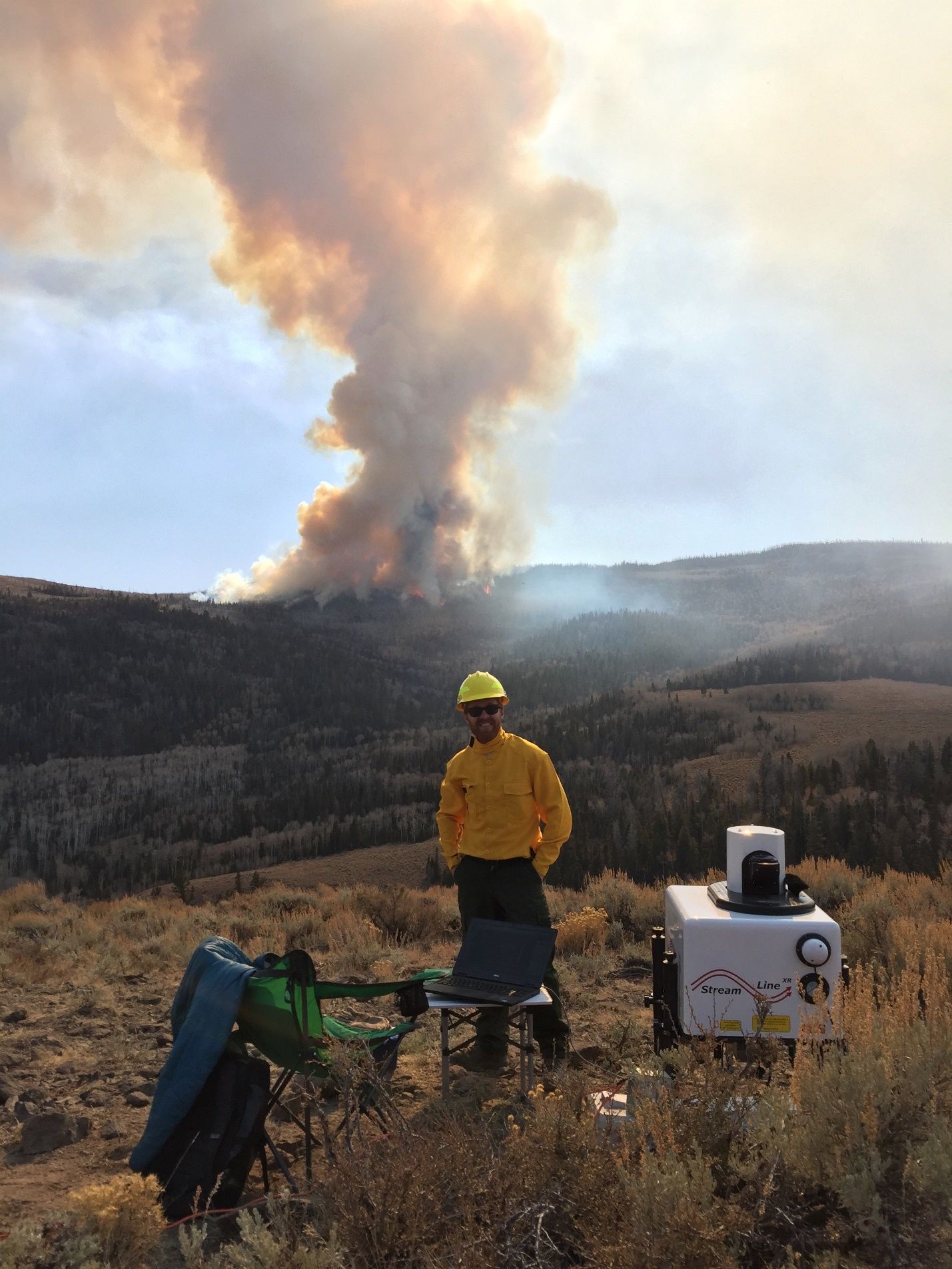 Lareau was at San Jose State University when he posed with a Doppler lidar a few kilometers from an active wildfire―the kind that creates its own clouds and weather. Photo is Courtesy of Neil Lareau. 