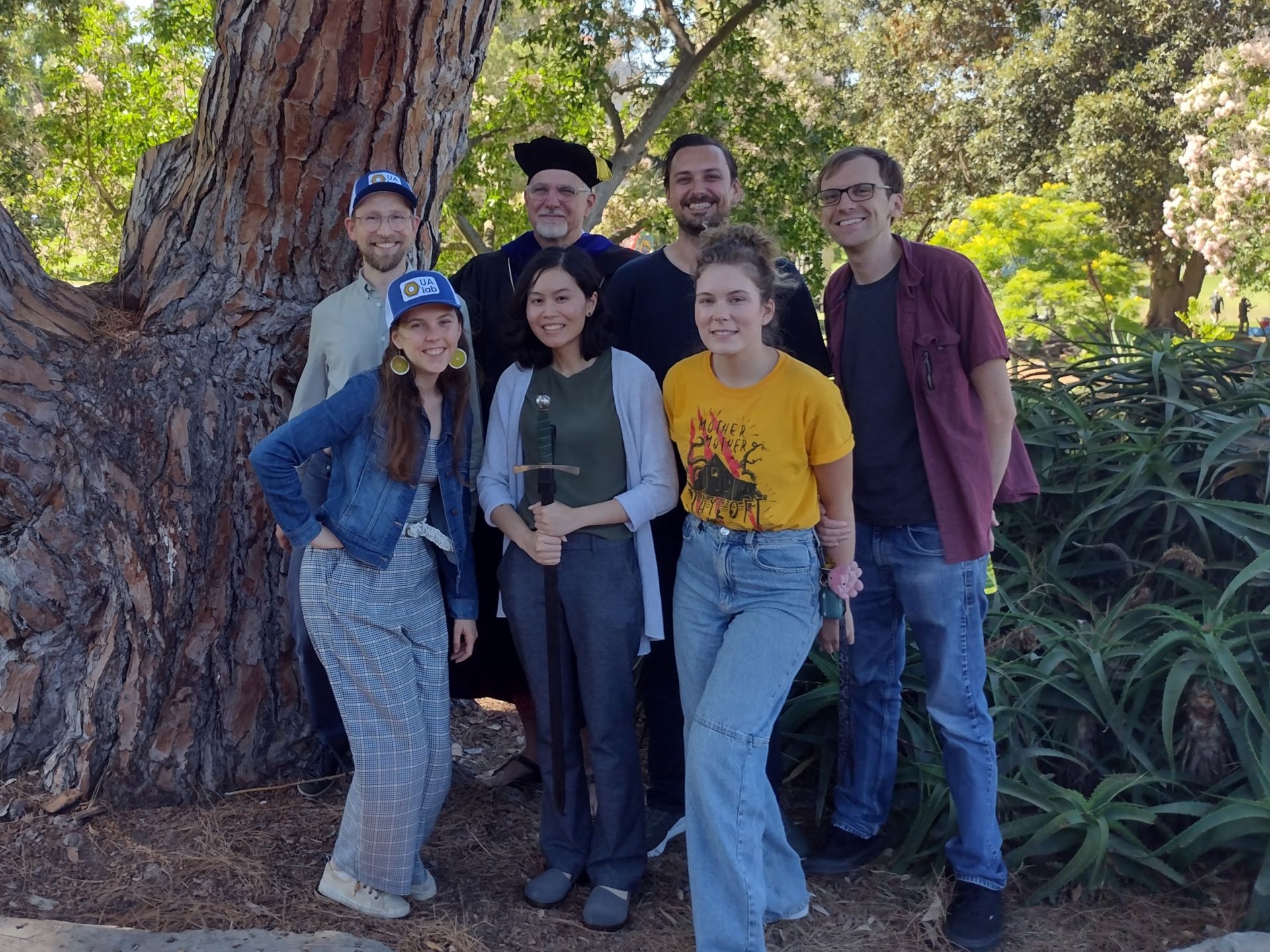 In July 2023, The Smith Ultrafine Aerosol Group paused for a portrait on the Irvine campus. From left, at rear, are postdoc Paulus Bauer, Smith, graduate student Jeremy Wakeen, and graduate student Adam Thomas. From left to right, in front, are graduate student Anna Kapp, freshly minted PhD Michelia Dam, and graduate student Kristen Cramer. The sword is engraved with the names of everyone who gets a PhD while part of the group, as Dam just did. Smith’s academic garb is part of the sword ceremony. Photo courtesy of Smith. 